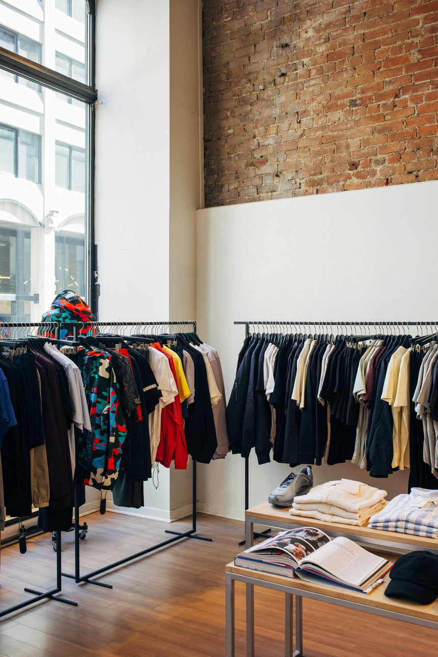 Clothing rack with various shirts and jackets, alongside a table displaying folded garments and magazines, set in a trendy retail space with exposed brick walls.