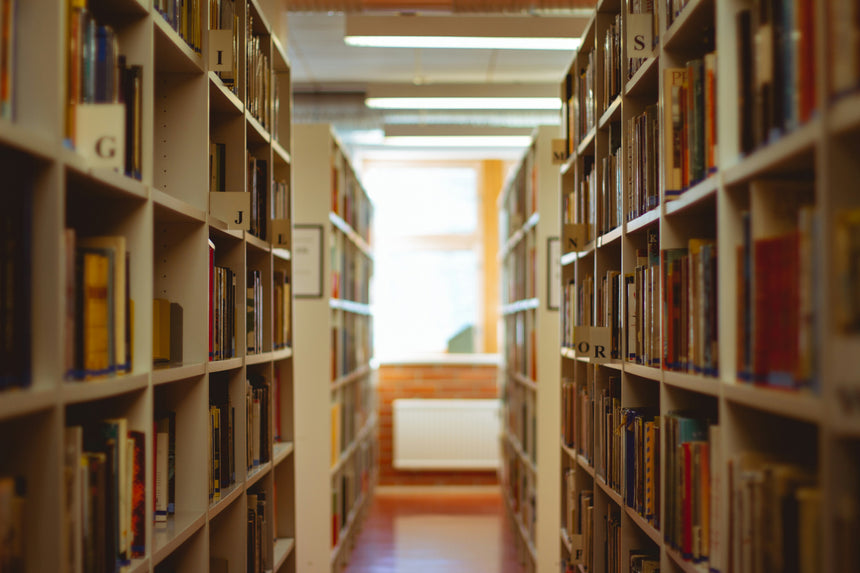 A library aisle with tall bookshelves on both sides, filled with books, leading toward a bright window in the distance.