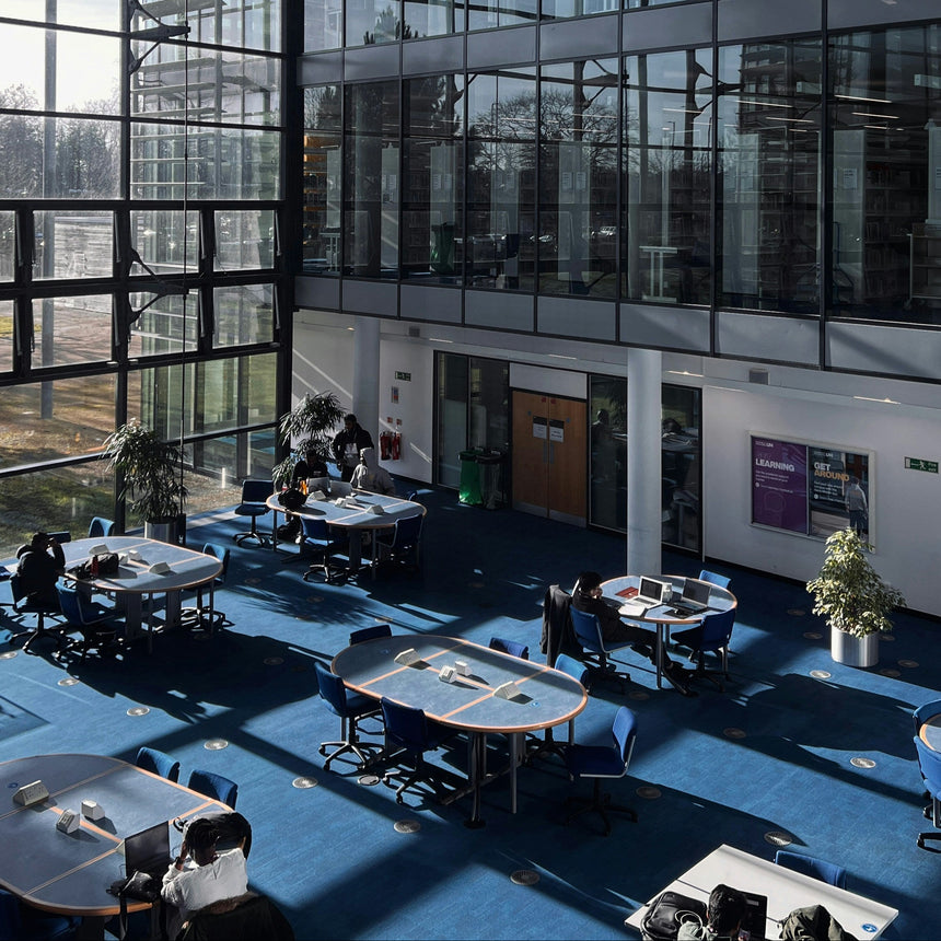 A large modern glass atrium interior with multiple study tables and chairs on a blue carpet, people seated and working, and tall windows surrounding the space.