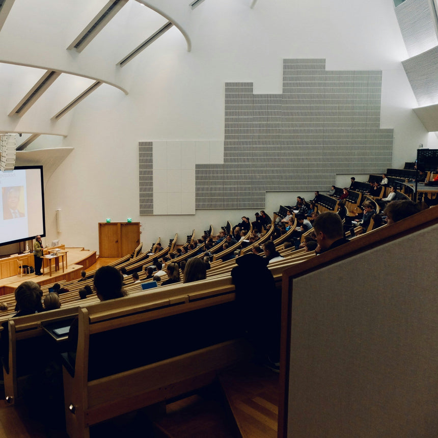 Large lecture hall with students seated and a presenter at the front, large screen displaying content.