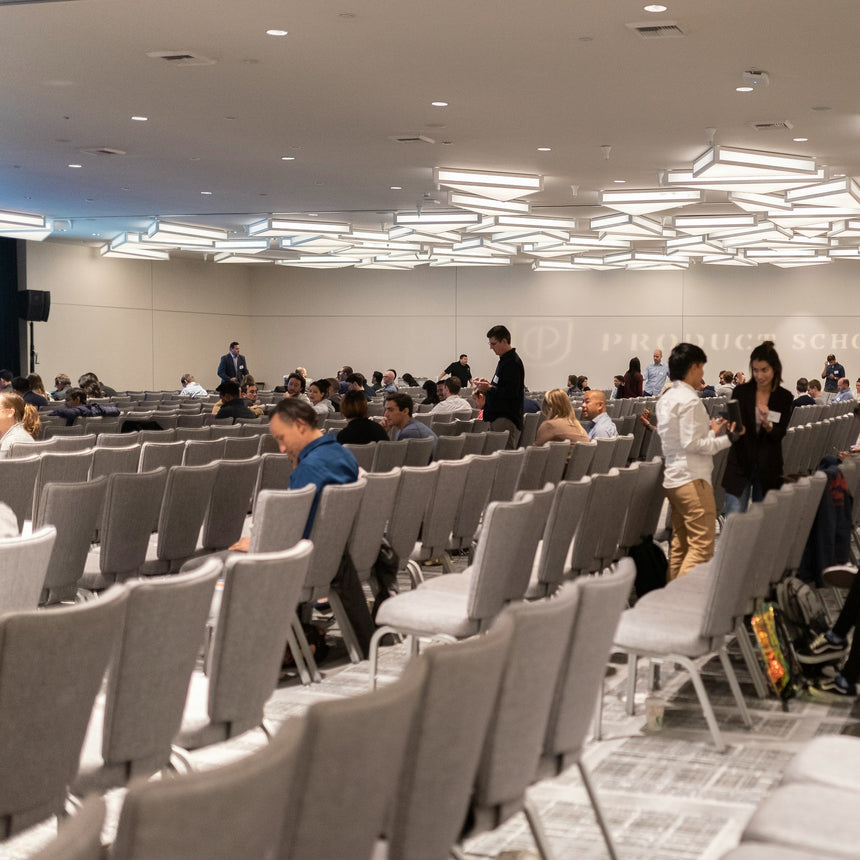 A large conference or seminar hall filled with rows of gray chairs, seated and standing people, and a stage area at the front under bright overhead lighting.