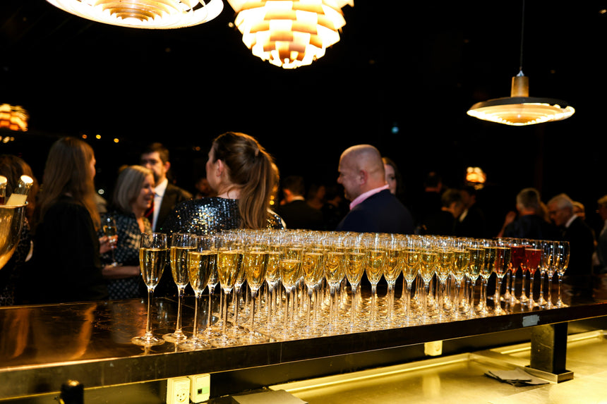 Rows of filled champagne glasses on a bar with people socializing in the background at a formal event, under warm hanging lights.