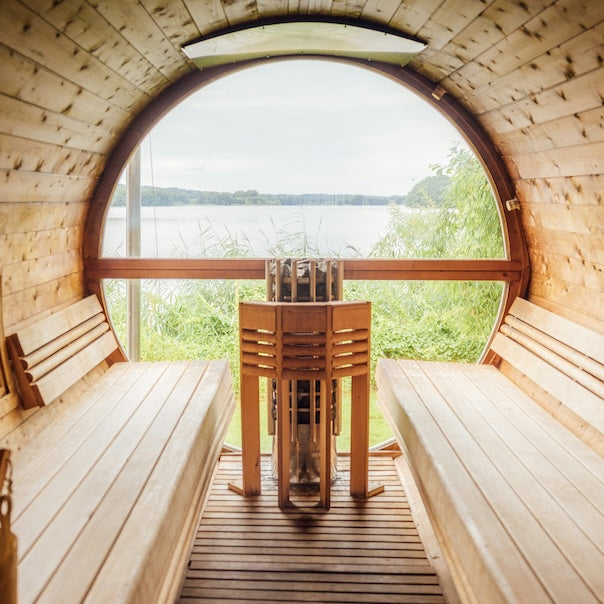 A wooden sauna interior with two long benches facing a large arched window overlooking a lake and green shoreline. A metal sauna stove sits centered on the wooden floor.