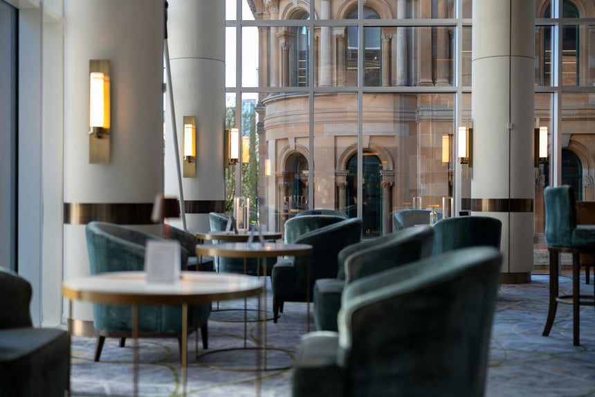 Modern lounge with plush teal chairs, round tables, and tall windows revealing a historic stone building outside. Warm wall lights illuminate the contemporary, relaxed setting.