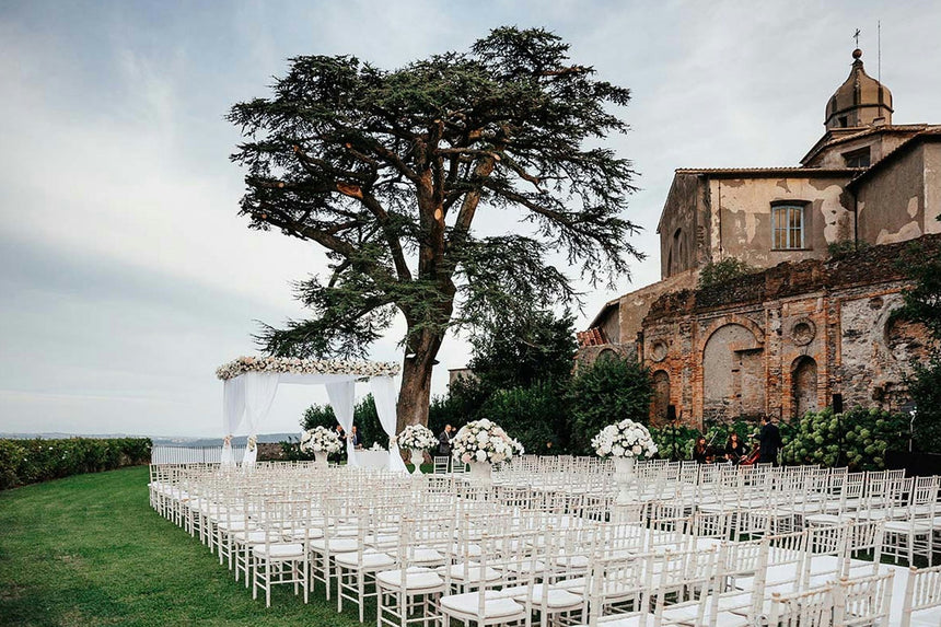 Elegant outdoor wedding ceremony with white seating and floral arrangements beneath a large tree