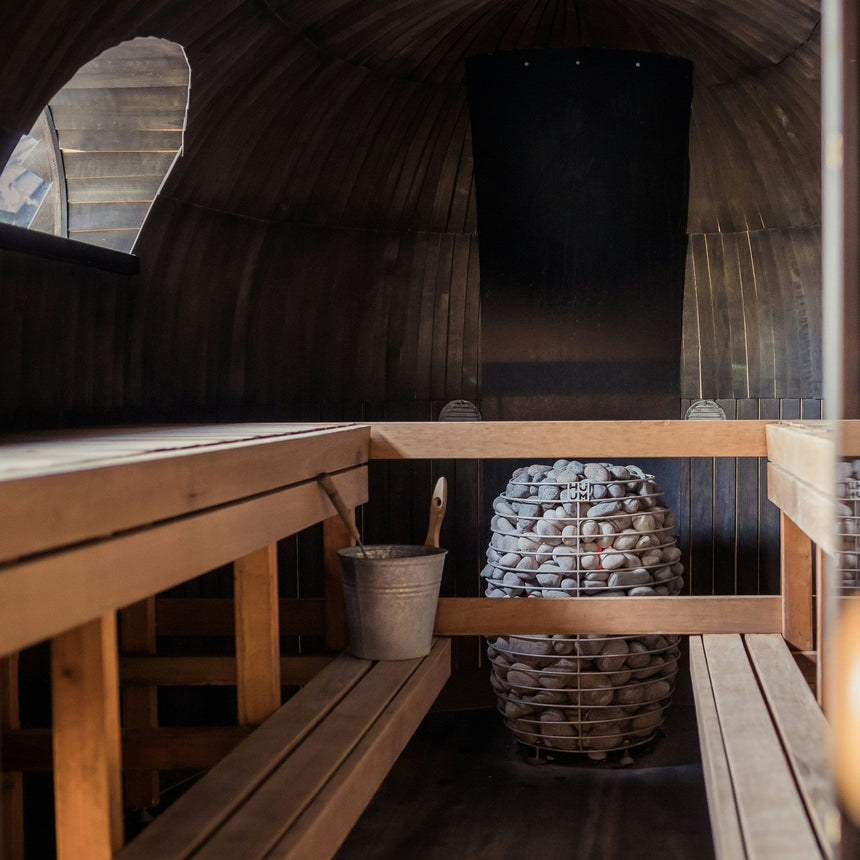Interior of a wooden sauna with benches on both sides, a bucket and ladle on the left, and a large pile of sauna stones in a metal holder at the center. Soft light filters in through a small window.