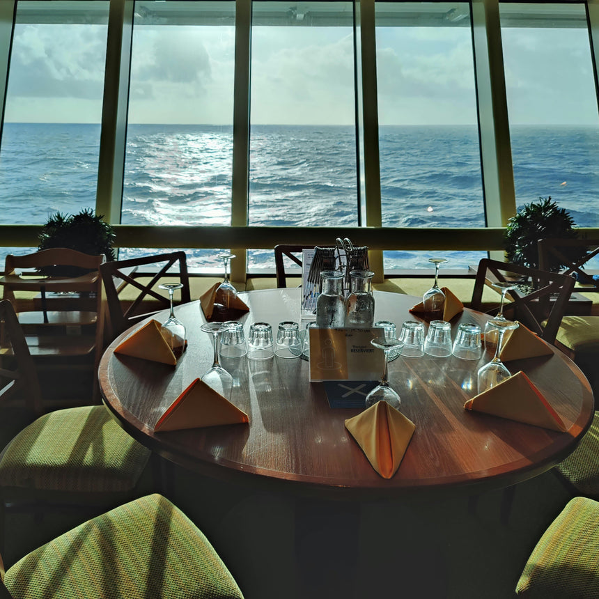 A round wooden dining table set with folded napkins, clear drinking glasses, and a small center display, surrounded by green upholstered chairs in a restaurant or bar with large windows overlooking the ocean.
