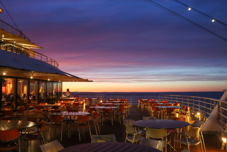Outdoor dining area on a cruise ship at sunset, with empty tables and chairs. The sky is a gradient of blue, purple, and orange, and string lights are overhead. The horizon and calm sea are visible in the background.