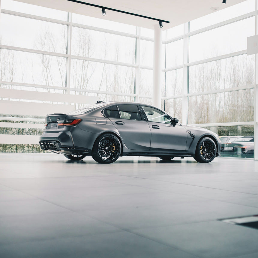 A matte gray BMW M3 sedan with black wheels, parked inside a modern, bright showroom with large windows.