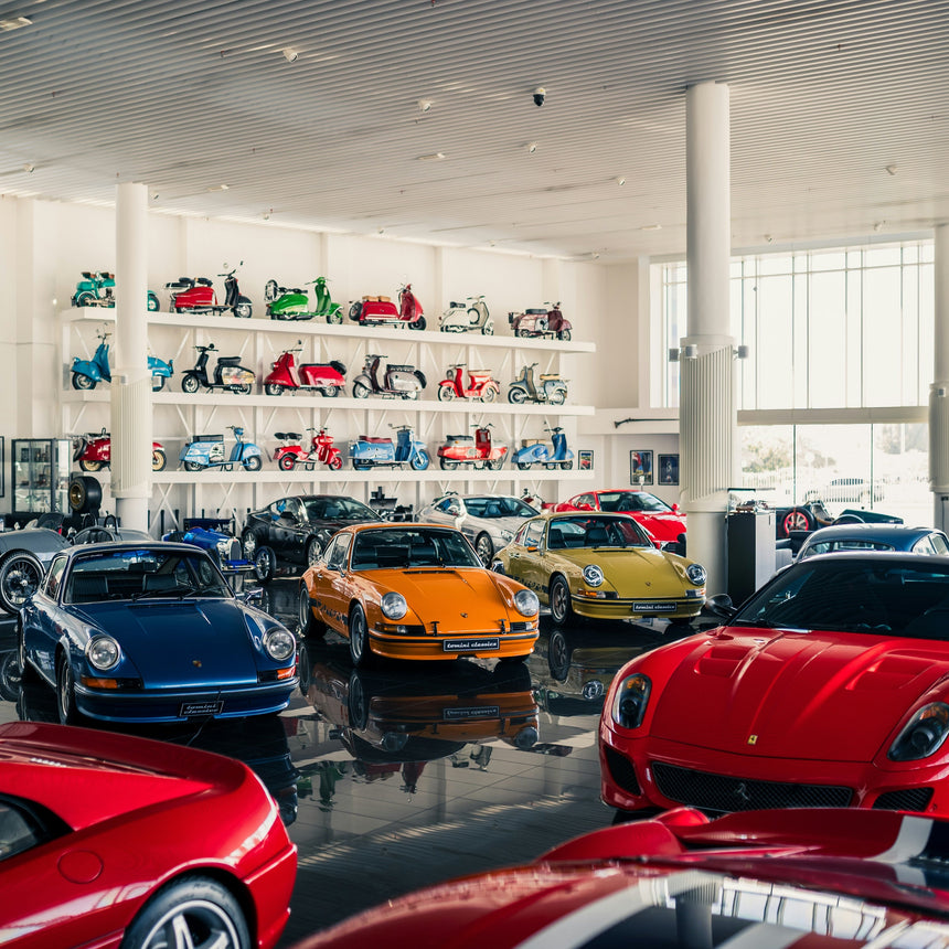 A luxury car showroom filled with classic sports cars, including a blue Porsche, an orange Porsche, a yellow Porsche, and a red Ferrari, with other cars displayed on white wall shelves in the background.
