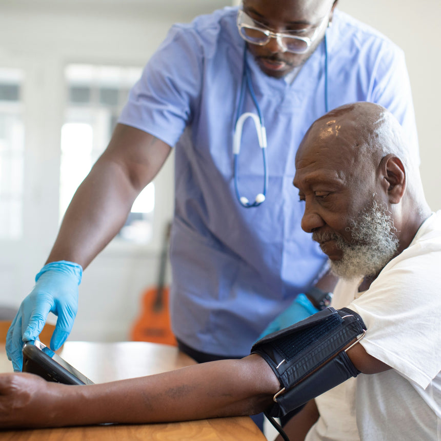 A healthcare worker in blue scrubs and gloves checks the blood pressure of an elderly man seated at a table. The man wears a white shirt and has a black blood pressure cuff on his upper arm in a bright indoor clinic setting.