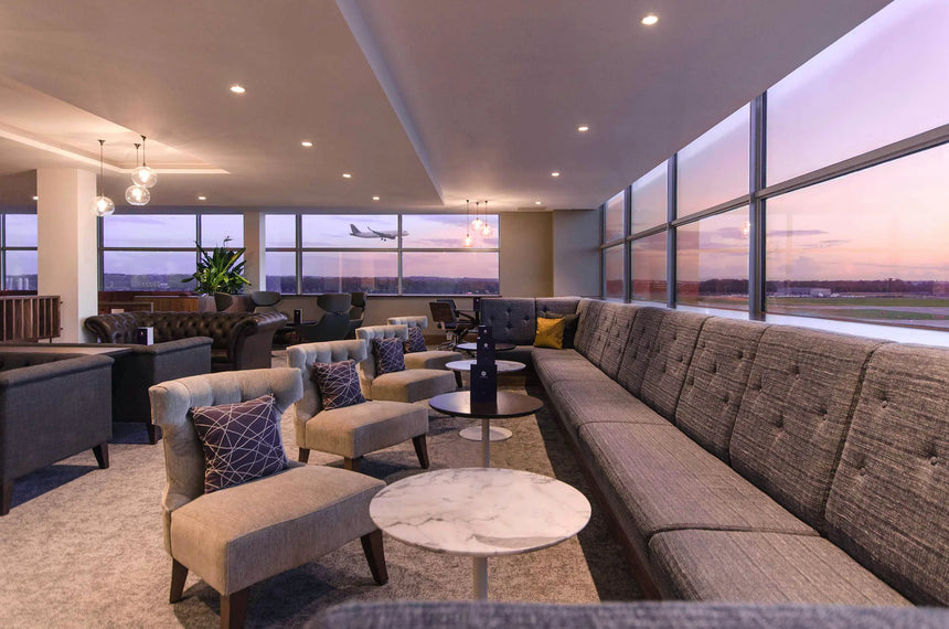 Modern airport lounge with gray sofas and armchairs, small round tables, large windows overlooking a runway, and a plane taking off at sunset. Soft lighting and a relaxed atmosphere.