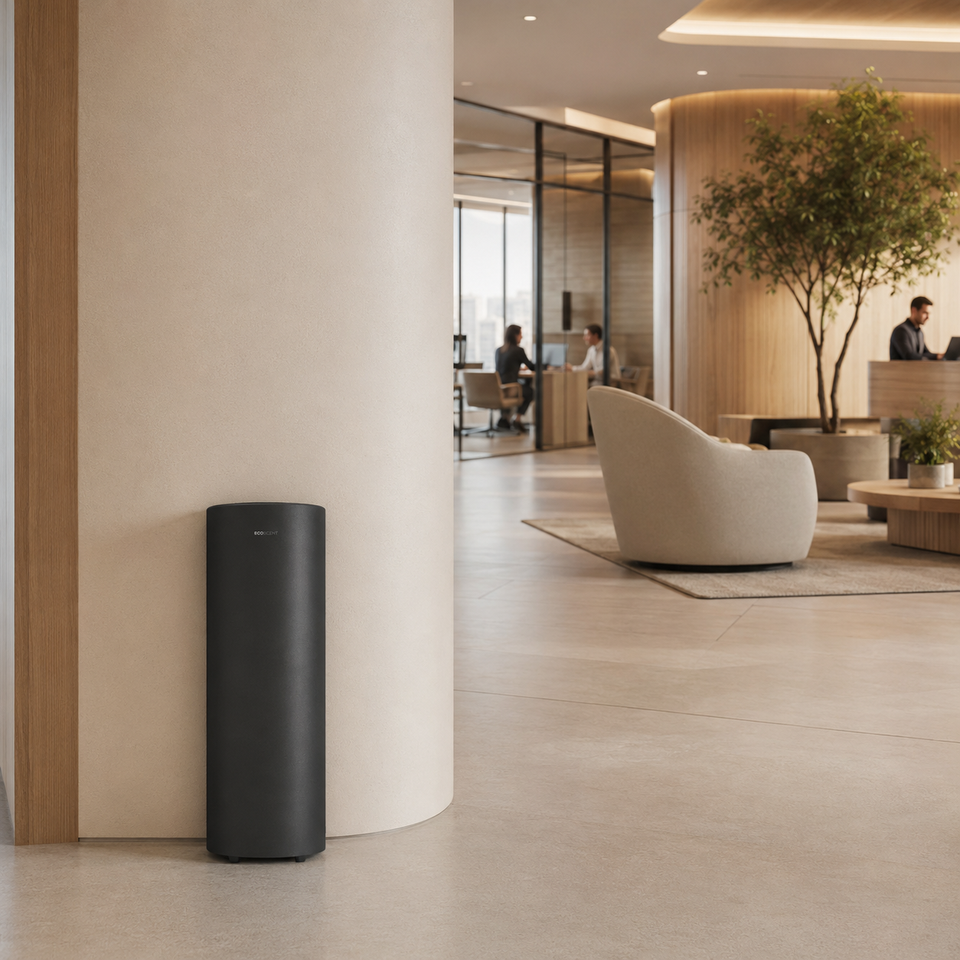 Black air purifier positioned near a column in a modern office space, with light-toned furniture and greenery visible in the background.