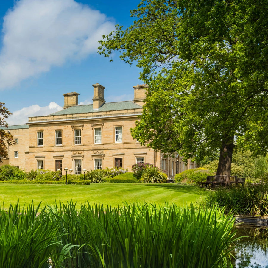 A grand beige stone mansion with a green roof, surrounded by a lush green lawn, trees, and a small pond under a blue sky with white clouds.