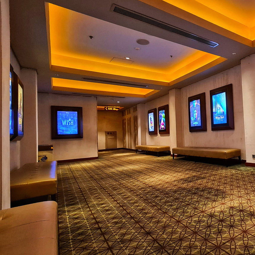 A wide interior lobby or event space with a patterned carpet, warm yellow recessed lighting, wall-mounted screens, and seating along the walls.