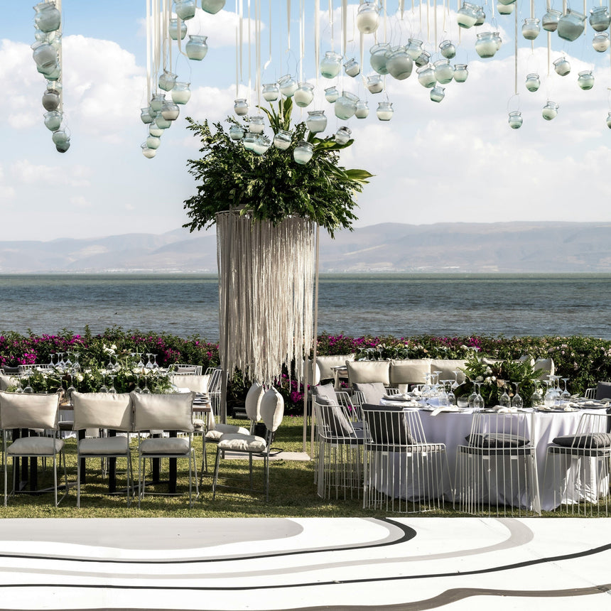 An outdoor event setup by the water with white draped tables, white chairs, two tall white draped pillars with green foliage, and numerous hanging white glass orb decorations suspended from above under a blue sky.