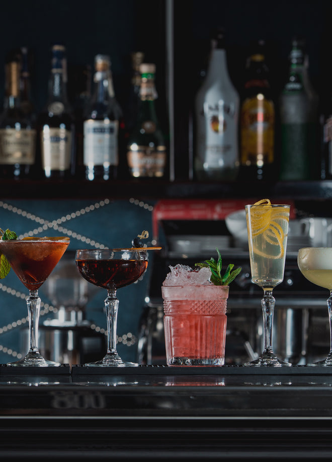 Ecoscent Bars & Nightclubs Signature Scents Samples set includes six 2ml vials, displayed among five cocktails in elegant glassware on a bar counter. The vials are situated on a polished surface with a dark backdrop.