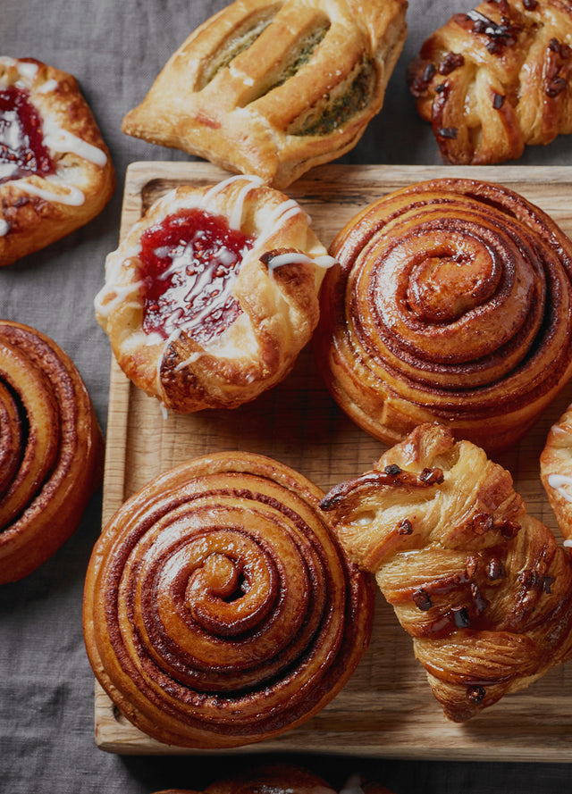 Ecoscent Food Signature Scents Samples 6 x 2ml featuring various pastries arranged on a wooden board, including cinnamon rolls and fruit-filled danishes, showcasing golden-brown crusts and glossy glazes.