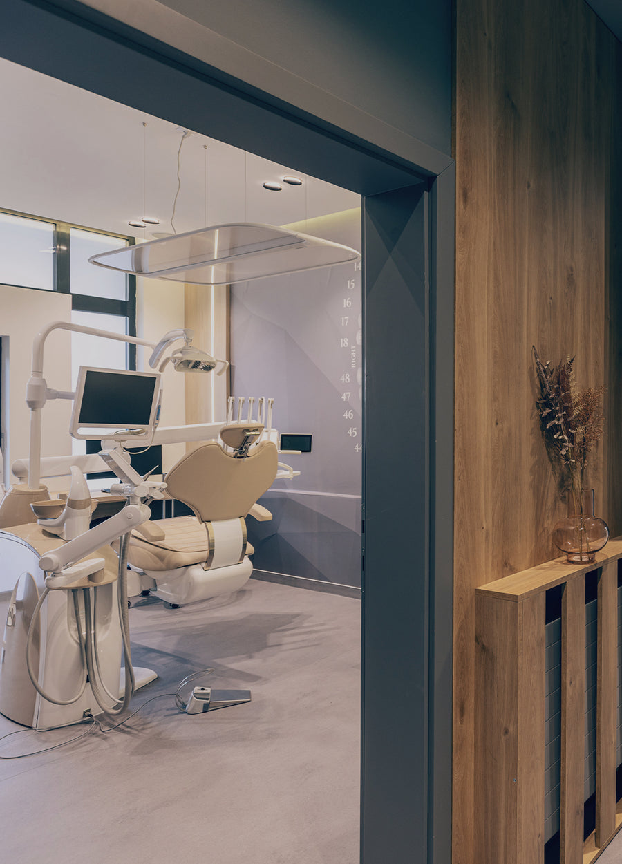 Dental treatment room with a reclining examination chair, a monitor, and dental equipment, seen through a doorway with a wooden partition.